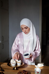 Muslim woman cooks dessert cupcake at home kitchen, arabian young model in white hijab and abaya. Islamic traditional clothing