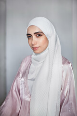 Portrait of young Muslim woman in white hijab headscarf and light pink abaya on wall background. Arab beautiful model