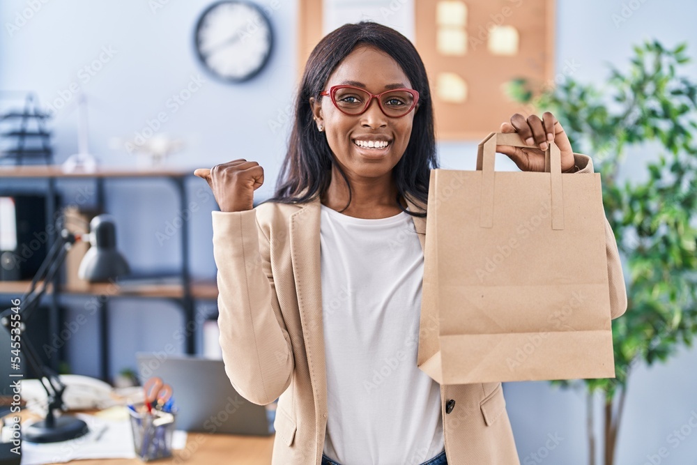 Sticker african young woman holding take away bag at the office pointing thumb up to the side smiling happy 