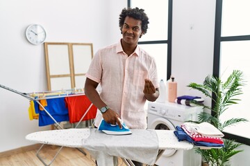 African man with curly hair ironing clothes at home doing money gesture with hands, asking for...