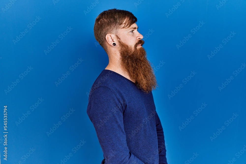 Canvas Prints Redhead man with long beard wearing casual blue sweater over blue background looking to side, relax profile pose with natural face and confident smile.