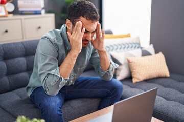 Young hispanic man stressed using laptop sitting on sofa at home