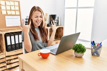 Young brunette woman working at the office with laptop shouting with crazy expression doing rock symbol with hands up. music star. heavy concept.