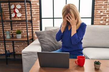 Middle age blonde woman using computer laptop at home with sad expression covering face with hands...