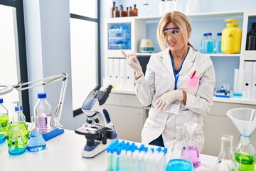 Middle age blonde woman working at scientist laboratory pointing aside worried and nervous with forefinger, concerned and surprised expression
