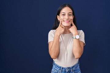 Young hispanic woman standing over blue background smiling with open mouth, fingers pointing and...