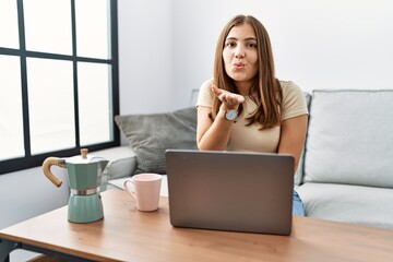 Young brunette woman using laptop at home drinking a cup of coffee looking at the camera blowing a...