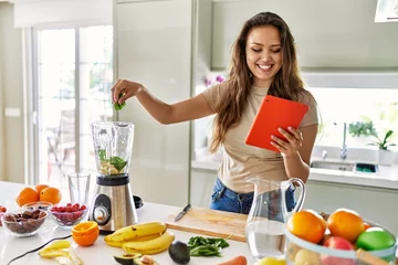 Tuinposter Young beautiful hispanic woman preparing smoothie with blender looking at recipe on tablet at the kitchen © Krakenimages.com