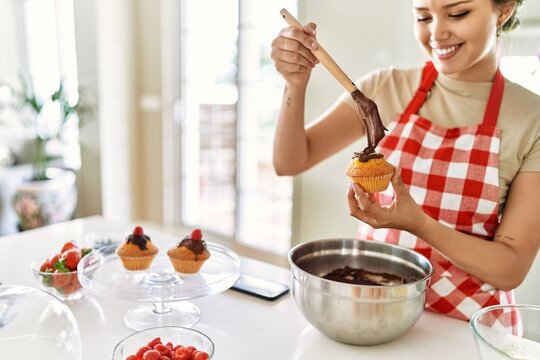 Young beautiful hispanic woman smiling confident putting chocolate on muffin at the kitchen