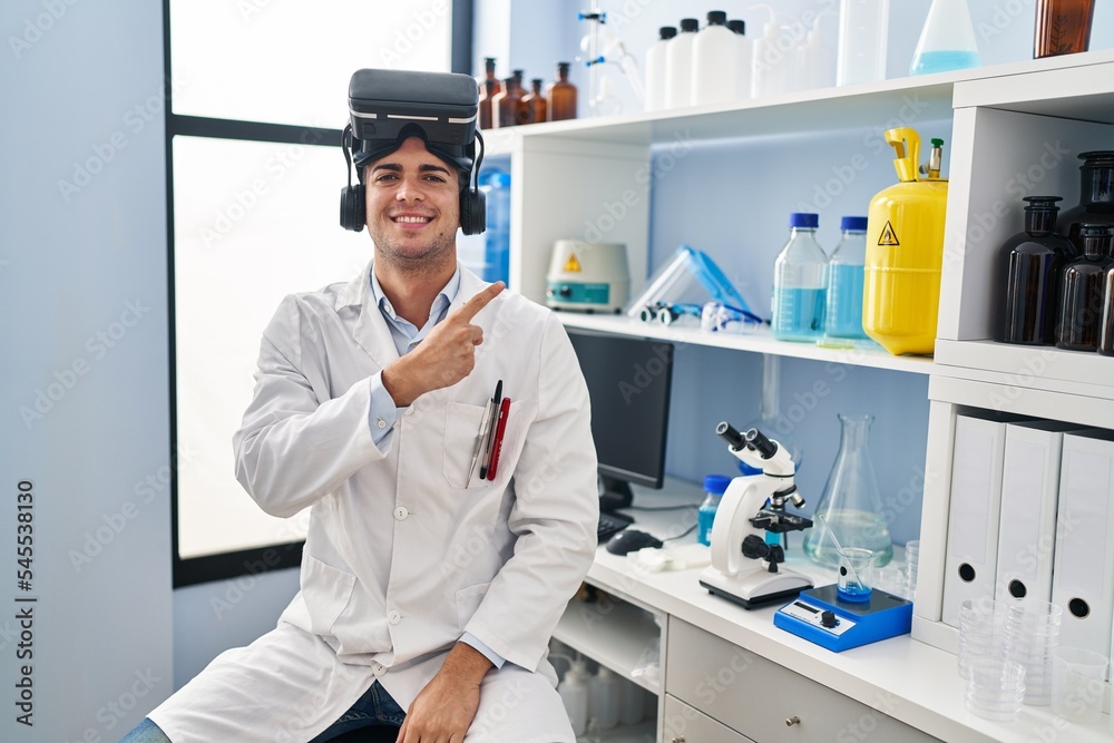 Wall mural Young hispanic man working at scientist laboratory wearing vr glasses smiling cheerful pointing with hand and finger up to the side