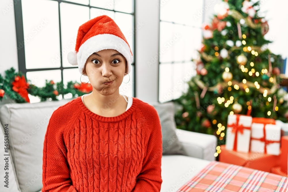 Sticker young hispanic woman with short hair wearing christmas hat sitting on the sofa puffing cheeks with f