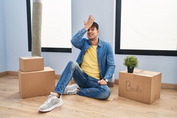 Young hispanic man sitting on the floor at new home surprised with hand on head for mistake,...