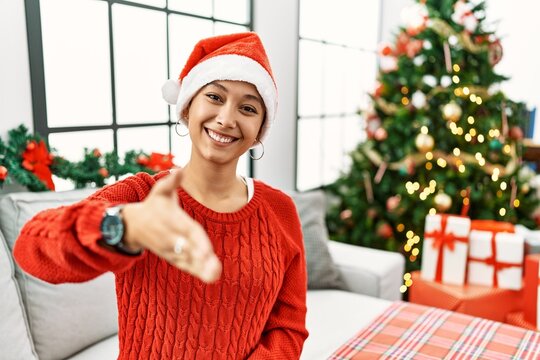 Young hispanic woman with short hair wearing christmas hat sitting on the sofa smiling friendly offering handshake as greeting and welcoming. successful business.