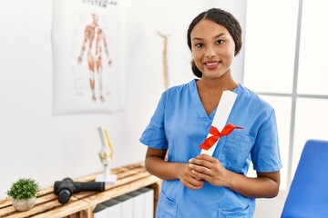 Young latin woman wearing physiotherapist uniform holding diploma at clinic