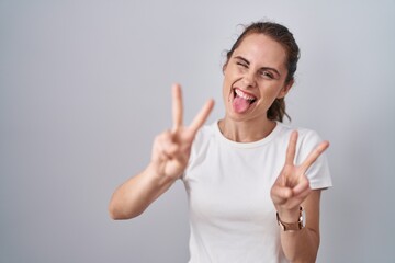 Beautiful brunette woman standing over isolated background smiling with tongue out showing fingers of both hands doing victory sign. number two.