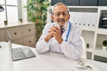 Senior grey-haired man doctor smiling confident sitting on table at clinic