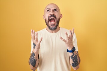 Hispanic man with tattoos standing over yellow background crazy and mad shouting and yelling with aggressive expression and arms raised. frustration concept.