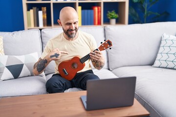 Young bald man having online ukulele class sitting on sofa at home