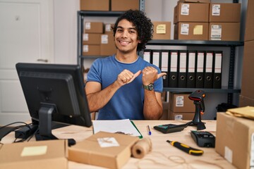 Hispanic man with curly hair working at small business ecommerce pointing to the back behind with hand and thumbs up, smiling confident