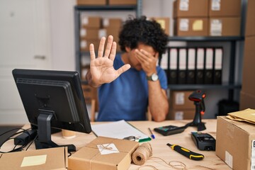 Hispanic man with curly hair working at small business ecommerce covering eyes with hands and doing...