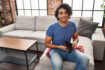 Young hispanic man showing empty wallet sitting on sofa at home
