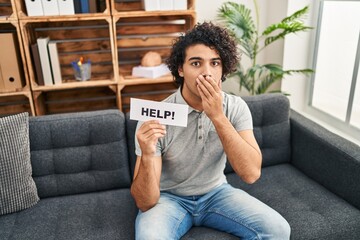 Hispanic man with curly hair asking for help covering mouth with hand, shocked and afraid for mistake. surprised expression