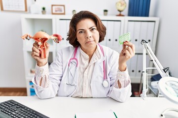 Middle age hispanic doctor woman holding anatomical female genital organ and birth control pills...