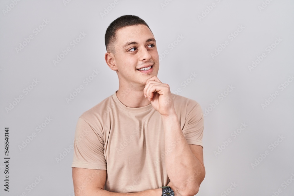 Wall mural Young man standing over isolated background with hand on chin thinking about question, pensive expression. smiling and thoughtful face. doubt concept.