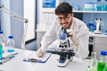 Young hispanic man scientist writing on document using microscope at laboratory