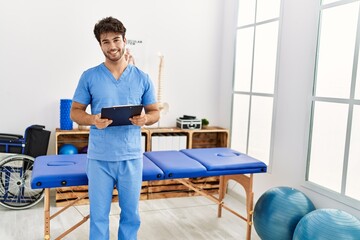 Young hispanic man wearing physio therapist uniform holding clipboard at clinic