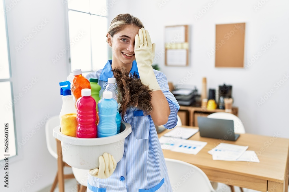 Sticker young blonde woman wearing cleaner uniform holding cleaning products covering one eye with hand, con