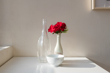 Bright red geranium flowers in white vase on table with bottle and bowl in sunlight with vintage...