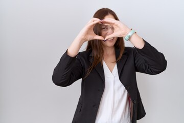 Beautiful brunette woman wearing business jacket and glasses doing heart shape with hand and fingers smiling looking through sign