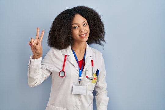 Young african american woman wearing doctor uniform and stethoscope smiling looking to the camera showing fingers doing victory sign. number two.