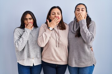 Mother and two daughters standing over blue background bored yawning tired covering mouth with hand. restless and sleepiness.