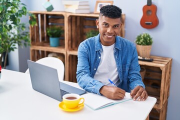 Young latin man using laptop writing on notebook at home