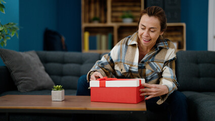 Middle age hispanic woman unboxing gift sitting on sofa at home