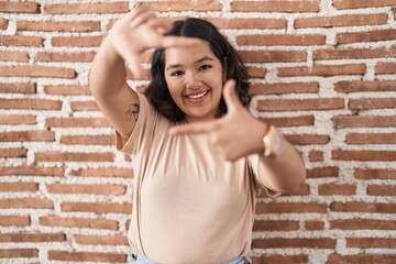 Young hispanic woman standing over bricks wall smiling making frame with hands and fingers with happy face. creativity and photography concept.