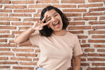 Young hispanic woman standing over bricks wall doing peace symbol with fingers over face, smiling cheerful showing victory