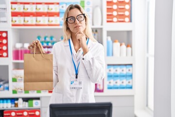 Young blonde woman working at pharmacy drugstore holding paper bag serious face thinking about question with hand on chin, thoughtful about confusing idea