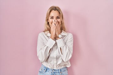 Young caucasian woman wearing casual white shirt over pink background laughing and embarrassed giggle covering mouth with hands, gossip and scandal concept