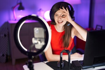Young caucasian woman playing video games recording with smartphone smiling happy doing ok sign with hand on eye looking through fingers