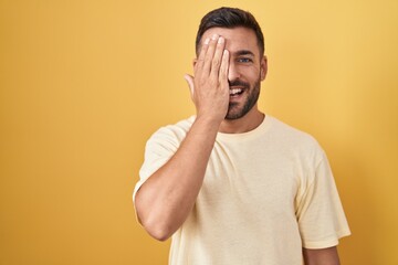 Handsome hispanic man standing over yellow background covering one eye with hand, confident smile on face and surprise emotion.