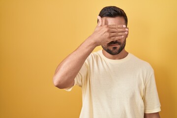 Handsome hispanic man standing over yellow background covering eyes with hand, looking serious and sad. sightless, hiding and rejection concept