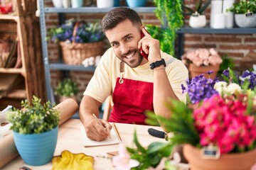 Young hispanic man florist talking on smartphone writing on notebook at florist