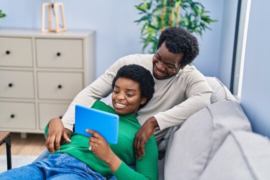 African american man and woman couple using touchpad sitting on sofa at home - Powered by Adobe
