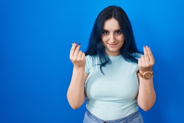 Young modern girl with blue hair standing over blue background doing money gesture with hands, asking for salary payment, millionaire business