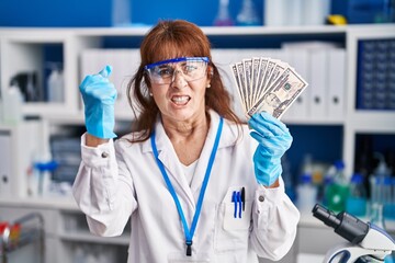 Middle age hispanic woman working at scientist laboratory holding dollars annoyed and frustrated shouting with anger, yelling crazy with anger and hand raised