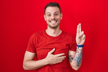 Young hispanic man standing over red background smiling swearing with hand on chest and fingers up,...
