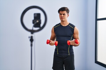 Hispanic man recording training tutorial at the gym skeptic and nervous, frowning upset because of...